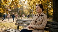 Middle-aged Caucasian woman in beige trench coat sitting on park bench during golden autumn day, smiling peacefully with warm sunlight filtering through trees.