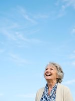 Senior woman with gray hair and fair complexion, wearing a cream cardigan over a blue floral blouse, smiling while looking upward against a bright blue sky with wispy clouds, outdoor portrait.