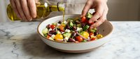 Fresh Mediterranean salad with olive oil dressing being prepared in modern kitchen. Healthy vegetarian meal with tomatoes, olives, cheese and herbs.