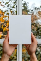 Woman holding blank white card with autumn foliage visible through window behind, ideal for greeting cards, invitations, marketing mockups and seasonal promotional designs.