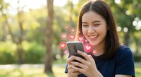 Happy young Asian woman using smartphone with floating heart icons in sunny park, social media engagement and digital communication concept.