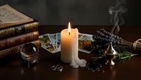 Mystical fortune telling setup with tarot cards, burning candle, crystal ball, dried herbs and antique books on dark wooden table for divination practices.