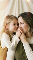 Young blonde daughter whispering secret to smiling mother in cozy sweaters sharing intimate bonding moment at home with warm natural lighting.