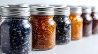 Glass mason jars filled with colorful dried fruits and berries arranged in row on white background, showing variety of preserved natural snacks and ingredients.