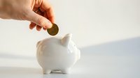 Hand inserting coin into white ceramic piggy bank on clean surface for savings and financial planning goals.