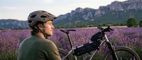 Young Caucasian man cyclist with helmet resting beside mountain bike in blooming lavender field at sunset with dramatic mountain backdrop for outdoor adventure.