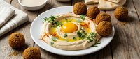 Creamy hummus topped with sunny side up eggs and fresh herbs served with golden falafel balls on rustic wooden table for Mediterranean breakfast or brunch meal.