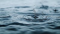 Water drops creating concentric ripple patterns on dark blue water surface with floating droplets captured in mid-air showing surface tension and wave motion.