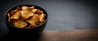 Crispy golden potato chips in black bowl on dark wooden table surface with dramatic lighting for snack food advertising and culinary photography.