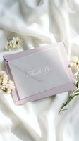 Elegant thank you card in a pastel lavender envelope surrounded by delicate white flowers on soft silk fabric, perfect for wedding stationery concepts.