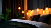 Cozy bedroom interior with glowing Edison bulb pendant lights creating warm atmosphere above bed with dark green bedding and decorative pillows.