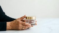 Mature woman with medium skin tone holding luxury skincare jar with gold lid on marble surface, showcasing cosmetics product for beauty and wellness marketing.