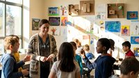 Female teacher engaging with diverse elementary students in colorful classroom with artwork displays and natural lighting from large windows.