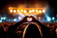 Silhouette of hands making heart shape at music concert with glowing lights and crowd in background, capturing love and celebration atmosphere.