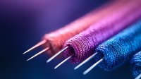 Vibrant macro shot of colorful sewing thread spools in pink, purple and blue tones arranged diagonally with metallic needles on dark background. Suitable for textile industry designs.