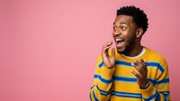 Excited young man with dark skin and short black hair wearing yellow and blue striped sweater, gesturing enthusiastically with both hands against pink background.
