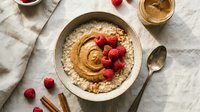 Creamy peanut butter hummus dip topped with fresh raspberries, served in a ceramic bowl with cinnamon sticks and spoon on marble surface for entertaining and snacking.
