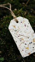 Handmade seed paper tag with embedded wildflower seeds and botanical materials, suspended by twine over lush green moss. Eco-friendly gift tag for sustainable packaging and gardening projects.