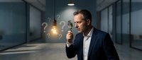 Middle-aged Caucasian businessman in dark suit holding glowing light bulb with floating question marks in modern office corridor representing innovation and problem-solving.