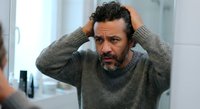 Stressed middle-aged Hispanic man with beard holding head in hands showing frustration and mental exhaustion in modern bathroom mirror reflection.