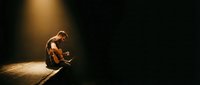 Young male musician sitting alone on stage playing acoustic guitar under dramatic spotlight with warm golden lighting against dark background.