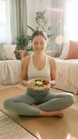 Young woman of mixed ethnicity in athletic wear sitting cross-legged on yoga mat holding bowl of healthy salad in bright bedroom with plants and natural light for wellness lifestyle content.