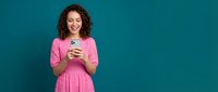 Young Hispanic woman in pink polka dot dress holding smartphone and smiling against teal background for social media and technology concepts.