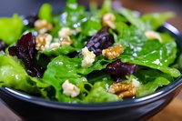 Fresh mixed green salad with walnuts and blue cheese crumbles served in dark ceramic bowl, closeup view of healthy organic lunch dish on wooden surface.