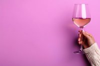 Female hand in beige sweater holding glass of rose wine against vibrant pink background, minimalist composition with copy space for wine tasting events.