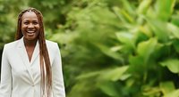 Young Black woman with long braided hair wearing white blazer smiling outdoors against lush green foliage background, professional portrait for business and corporate use.