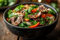Asian noodle bowl with sliced beef, red bell peppers, fresh herbs and vegetables served in dark ceramic bowl on wooden table, close up view.