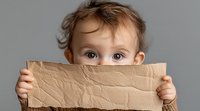 Young child with big expressive eyes peeks over wrinkled kraft paper held up to cover lower face, creating playful hide and seek moment against neutral background.