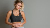Young Caucasian woman with blonde hair experiencing stomach pain, wearing gray sports bra against neutral background, expressing discomfort with hand on abdomen.