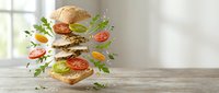 Stacked sandwich with flying fresh vegetables and ingredients on wooden surface near window, suitable for food advertising and restaurant marketing.