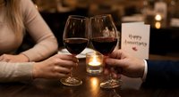 Couple toasting with red wine glasses at romantic dinner celebrating anniversary with candle and greeting card in cozy restaurant atmosphere.