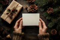 Hands holding blank card with Christmas gift, pine cones and fir branches on rustic wooden background. Cozy winter holiday flat lay composition.