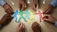 Diverse hands of multiple ethnicities arranging colorful paper cutout figures in circle on wooden surface, symbolizing teamwork, unity, community support and collaborative partnership for business concepts.
