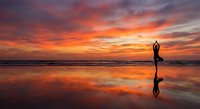 Silhouette of person practicing yoga tree pose on beach at sunset with dramatic orange sky reflected in wet sand creating mirror effect on ocean shore.