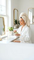 Young woman in white bathrobe and towel applying face mask in modern bathroom with gold mirrors and plants for skincare routine and self-care wellness.