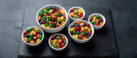 Colorful candy-coated chocolates in white ceramic bowls arranged on dark slate surface for party treats and confectionery displays.
