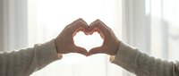 Hands forming a heart shape against a bright window with soft light, symbolizing love, care, and connection in a cozy home setting.