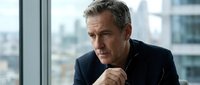 Mature male professional with gray hair and Caucasian ethnicity wearing dark business suit, sitting by floor-to-ceiling window in high-rise office with city skyline view, hand near chin in thoughtful pose, suitable for corporate, leadership, and business concepts.