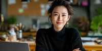 Confident Asian woman entrepreneur in black turtleneck sweater smiling at camera in cozy home office with laptop and plants.