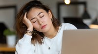 Exhausted Asian woman sleeping at desk with laptop, showing burnout and fatigue during remote work from home office.