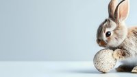 Adorable brown and white baby rabbit with spotted Easter egg on minimalist light background, perfect for holiday greeting cards and spring designs.