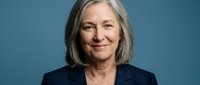 Senior woman with gray hair wearing dark blazer against blue background, professional headshot for business and corporate use.
