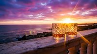 Two crystal whiskey glasses on wooden deck overlooking ocean at sunset with dramatic pink and orange sky creating romantic atmosphere for luxury dining.
