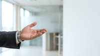 Caucasian male professional in business attire extending open palm gesture in modern office environment with glass partitions and natural light.