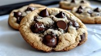 Fresh baked chocolate chip cookie with melted dark morsels on parchment paper, close up view showing soft, chewy texture and golden brown edges.