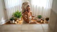 Young woman wrapped in towel enjoys spa day at home, talking on phone surrounded by candles and plants, skincare self-care routine.